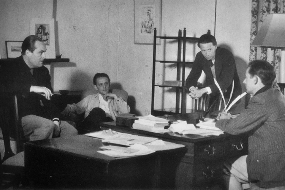 An old photo of a group of men in casual suits, surrounding a wooden table, chatting with each other.